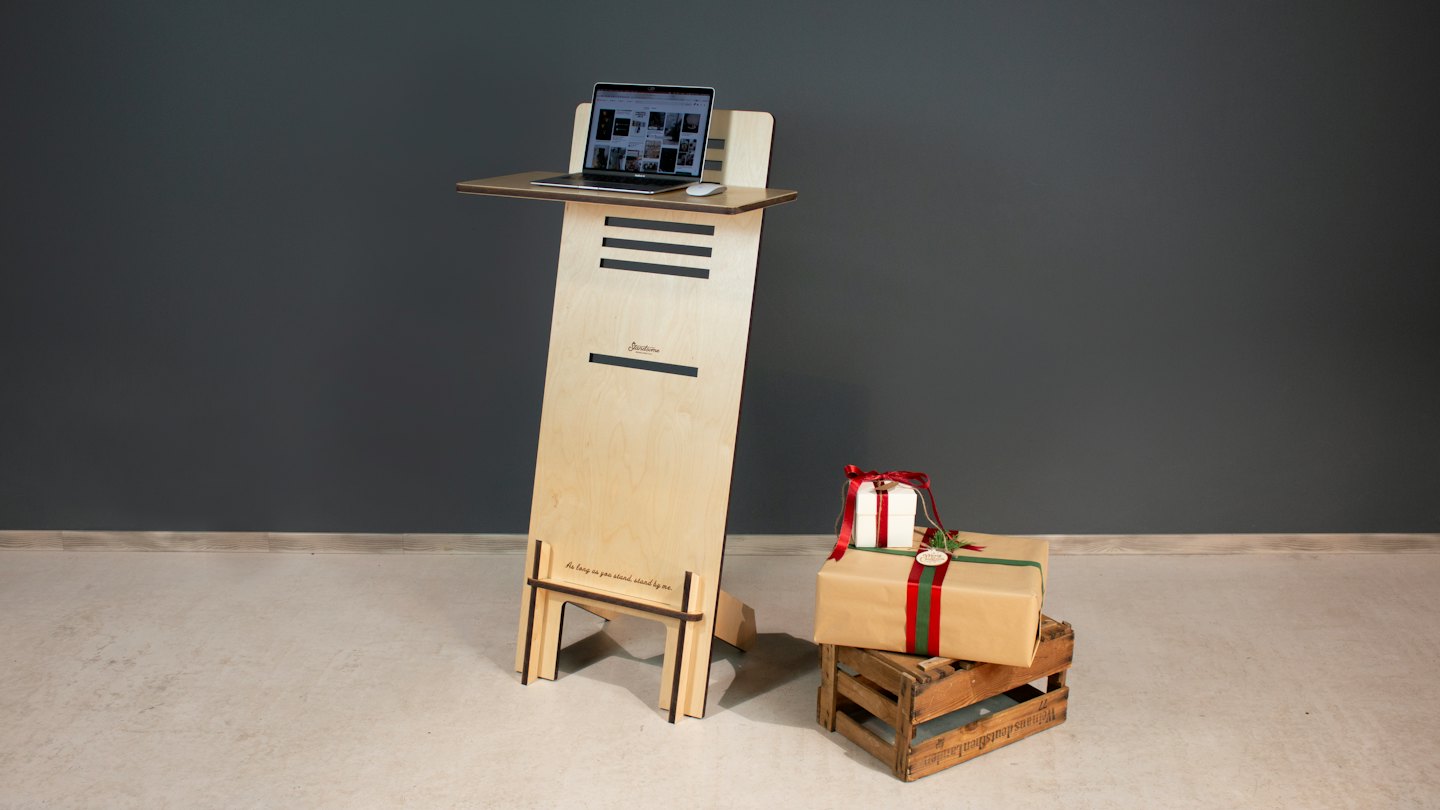 Wooden standing desk with laptop and mouse on a grey wall.